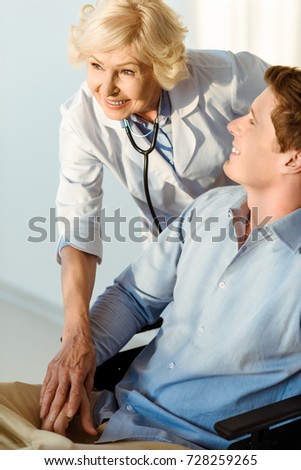 Smiling young man sitting in wheelchair with doctor standing over him, holding his hand and smiling