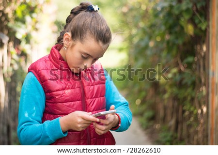 Beautiful girl is typing on a smartphone in  red vest outdoor.Handsome smiling girl with mobile phone walking on outdoor. Portrait smiles girl. copy space. The girl with a smartphone in the hands