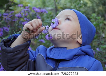 A little boy in blue demi-season clothing emotionally smells flowers looking up at the sky. A kid in a blue autumn jacket and hat looks dreamily and joyfully upwards into the future. 
