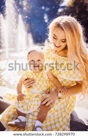 Beautiful blonde mother in yellow spotted dress sits with her daughter in the rays of morning sun