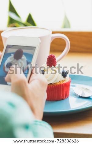 Woman takes a picture of cup with cappuccino and a cupcake on the plate, soft focus background
