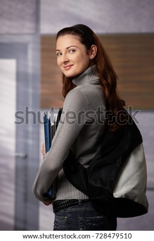 Happy female student standing on school corridor, holding workbooks, looking back over shoulder, smiling.