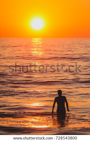 A vertical picture with a silhouette of a man walking in the sea towards the sunset.