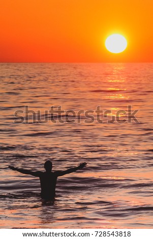 A vertical picture with silhouette of a man raising his arms while walking in the sea towards the sunset.