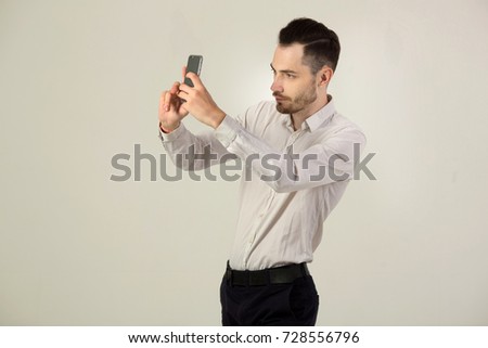 young brunette man in white shirt with unshaven face on background of light wall with phone in hand