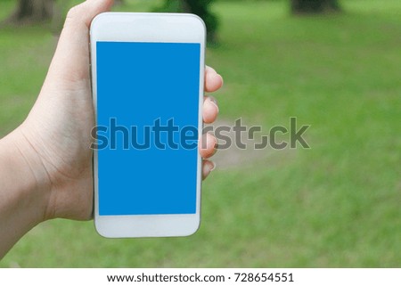 Left hand holding mobil phone on green grass and tree background. Blue screen smartphone for Graphic, Product, Application and Mock up display montage.