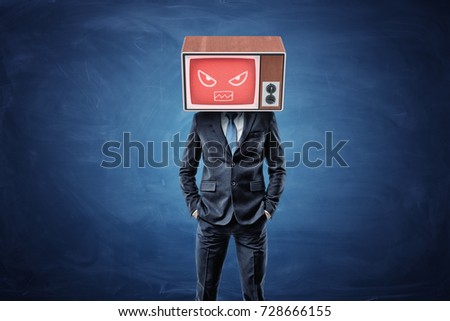 A businessman stands with hands in his pockets and wears a TV box on his head with a red screen showing an angry face. Business and success. Proactive approach. Fight off your competition.
