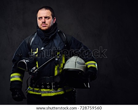 Studio portrait of firefighter dressed in uniform holds safety helmet in his arm.