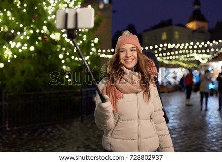holidays and people concept - beautiful happy young woman taking picture by selfie stick at christmas tree and market in winter evening