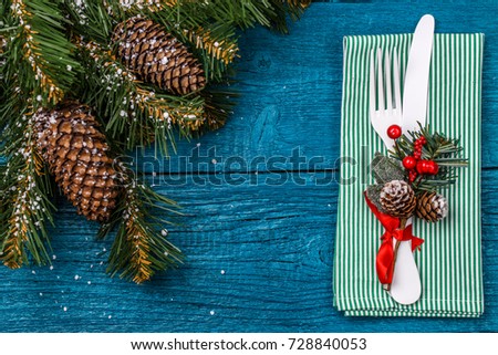 Christmas table place setting - blue table with green napkin, white fork and knife, decorated sprig of mistletoe and christmas pine branches. Christmas holidays background.