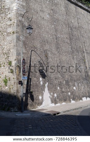 Squiggly street lamp with traffic signs, signpost and pedestrian light and its attached shadow casted onto the historical fortification wall at driveway to Castle Blois; Blois, France; 01/06/2017
