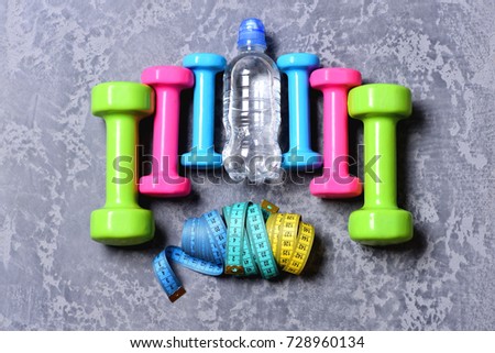Barbells, colorful tape measures and water bottle placed in pattern, top view. Shaping and fitness. Workout and sport concept. Dumbbells made of pink, green and cyan plastic on grey texture background