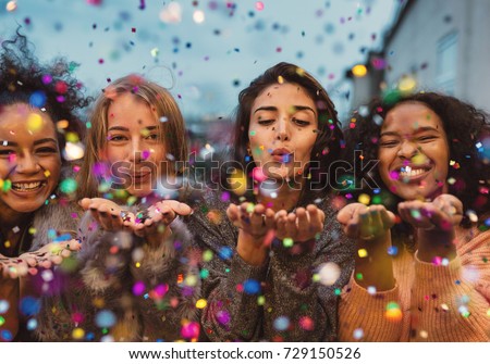 Young women blowing confetti from hands. Friends celebrating outdoors in evening at a terrace. Royalty-Free Stock Photo #729150526
