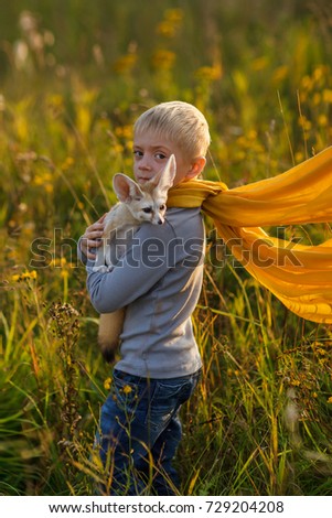 A boy in the image of the Little Prince Antoine Exupery holds a fox in his hands. Fennec tame. A child in a yellow scarf  look at the camera. Fox. Fennecus zerda. 