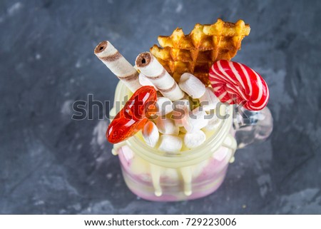 Freakshake from pink smoothie, cream. Monstershake with lollipops, waffles and marshmallow. Extreme milkshake in a Mason jar. Gray dark concrete background. Top view.