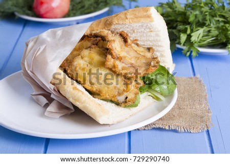 Deep Fried Fillet Sardines Fish Sandwich serving with round plate, green arugula salad, juicy lemon and sliced white onion on rustic blue background. Balik ekmek is Turkish Culture for cheap fast food