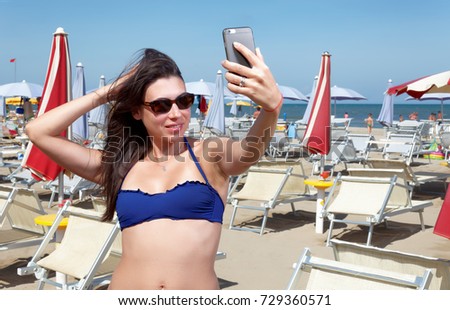 Young Happy Smiling dark hair girl / woman in blue bikini taking fun selfie portrait picture on beach vacation with red umbrellas and loungers - Cervia - Adriati sea - Emilia Romagna - Italy