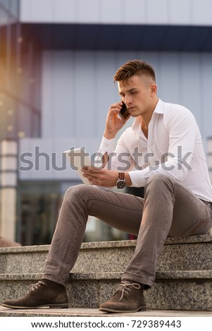 Serious businessman sitting on the staircase outdoors and talking on mobile phone while using digital tablet