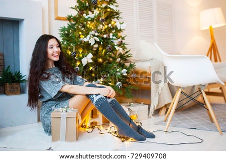 Beautiful girl in a gray dress with a gift in hand sits near a Christmas tree