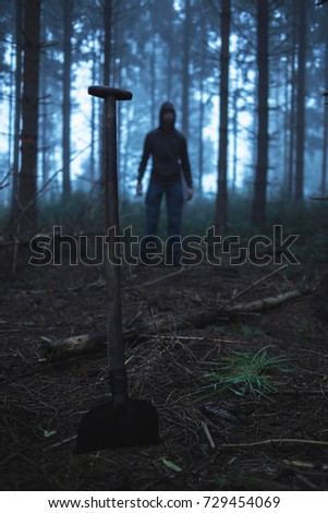 Shovel and man in hoody in spooky misty pine forest.