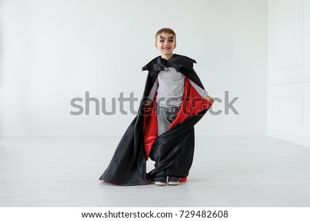 Playing boy with Halloween vampire makeup is fluttering his red, black cloak with bat like collar, in a light space with white ghosts on background. Smiling kid waiting halloween, copy space available