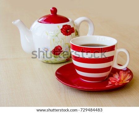 striped cup with tea on a saucer and brewer with a poppy picture on a wooden table top and red rose flower