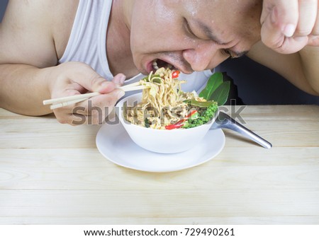 A man face full of sweat eating stir fried hot and spicy instant noodles in a white cup on wooden table