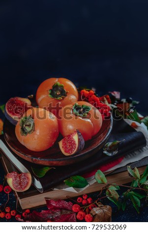 Dark autumn still life, ripe persimmons on a wooden plate with autumn leaves. Seasonal fruits close-up. Dark food photography with copy space.
