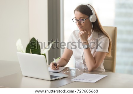 Focused attentive woman in headphones sits at desk with laptop, looks at screen, makes notes, learns foreign language in internet, online study course, self-education on web, consults client by video Royalty-Free Stock Photo #729605668