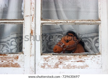Brown toy horse is looking for freedom out of an old glass window with wood frames in front of a retro handiwork white curtain