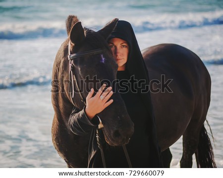 Young Blonde woman stroking and hugging horse. Beautiful lady with her black stallion on seashore enjoying nature. Love and friendship concept. Waves ocean background