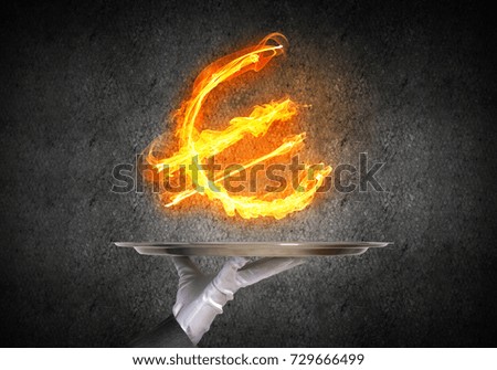 Cropped image of waitress's hand in white glove presenting flaming euro symbol on metal tray with dark wall on background.