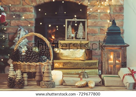 Dark Christmas composition, lantern, presents and handmade wooden decorations on the table in front of fireplace with woodburner, ornaments and garlands on the back, lights, selective focus, toned