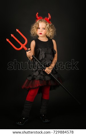 Vertical picture, isolated on black, attractive caucasian blonde little girl in black dress, dark boots and red tights, with red horns on head hold trident with both hands