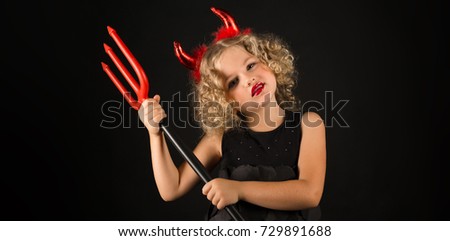 Widescreen picture, isolated on black, adorable caucasian blonde little girl in black dress, with red horns on head hold trident both hands, look at camera