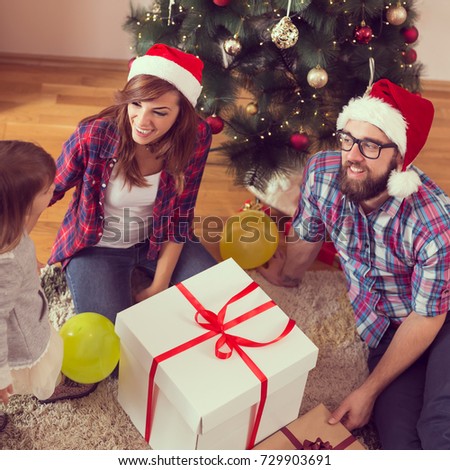 Young family sitting next to a nicely decorated Christmas tree, wearing Santa's hats and opening their presents