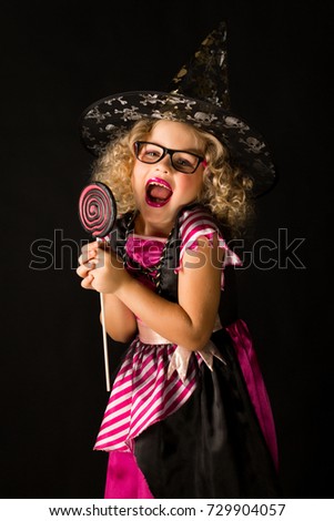 Vertical picture, isolated on black, cute caucasian blonde little girl in black dress with pink stripes, black scull hat and glasses holds big lollipop