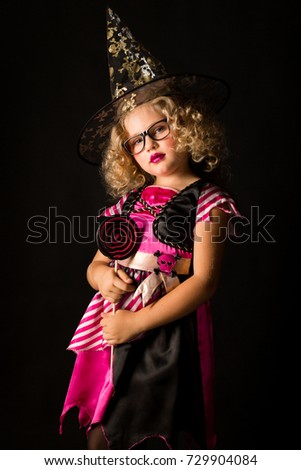 Vertical picture, isolated on black, cute caucasian blonde little girl in black dress with pink stripes, black scull hat and glasses holds big lollipop, look at camera