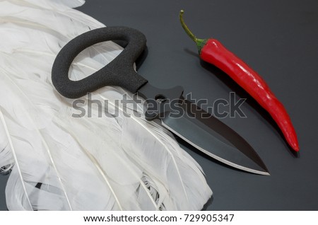 Black knife on a black background. White feathers and red pepper. Tactical concept.