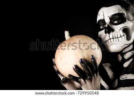 Isolated on black, closeup picture, young brunette caucasian pretty woman with scull body art with a pumpkin in hands, grey eyes, position at right