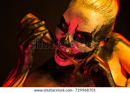 Isolated on black, closeup picture, toned red and yellow, beautiful young blonde caucasian woman with scull body art with wine glass in teeth, grey eyes, look at camera from brow