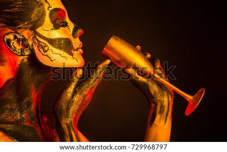 Isolated on black, closeup picture, toned red and yellow, beautiful young blonde caucasian woman with scull body art with wine glass in hand, grey eyes, drinks from glass with pleasure