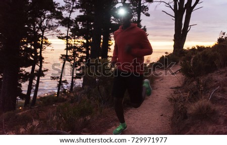Young African man wearing a headlamp running alone down a trail in the forest while out for a cross country run at dusk Royalty-Free Stock Photo #729971977