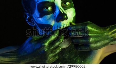 Isolated on black, wide-screen closeup picture, toned green and blue, spooky young blonde caucasian pretty woman with scull body art, grey eyes, both hands in her mouth, look at camera