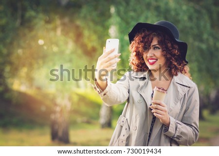 Beautiful young redhead woman taking a selfie in park in autumn. Closeup portrait of cute curly hair female person smiling taking a photo of herself outdoors. Retouched, vibrant colors, copy space.