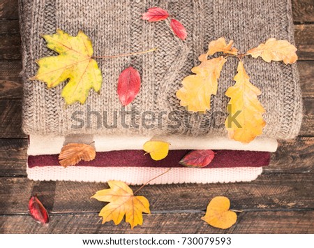 Pile of knitted winter clothes on wooden background covered with autumn leaves, knitwear. Stack of knitted sweaters and cardigans.