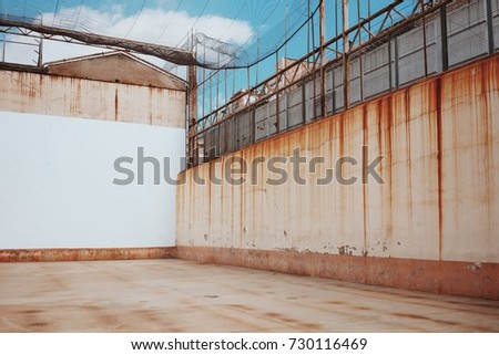 Empty backyard of old prison with rusty streaks on walls, grid and barbed wire fence under blue sky with clouds.