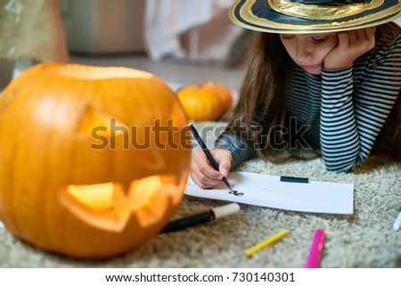 Portrait of pretty little girl wearing Halloween costume drawing laying on floor by carved pumpkin