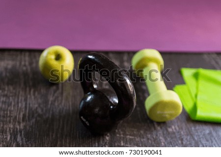 Closeup front view shot of a kettlebell, dumbbell, fitness gum and an apple aside exercise mat on a dark wooden floor background