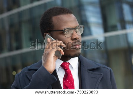 Urban closeup portrait of handsome African American executive standing in street with skyscrapers in background, while talking on cellphone discussing difficult business matters